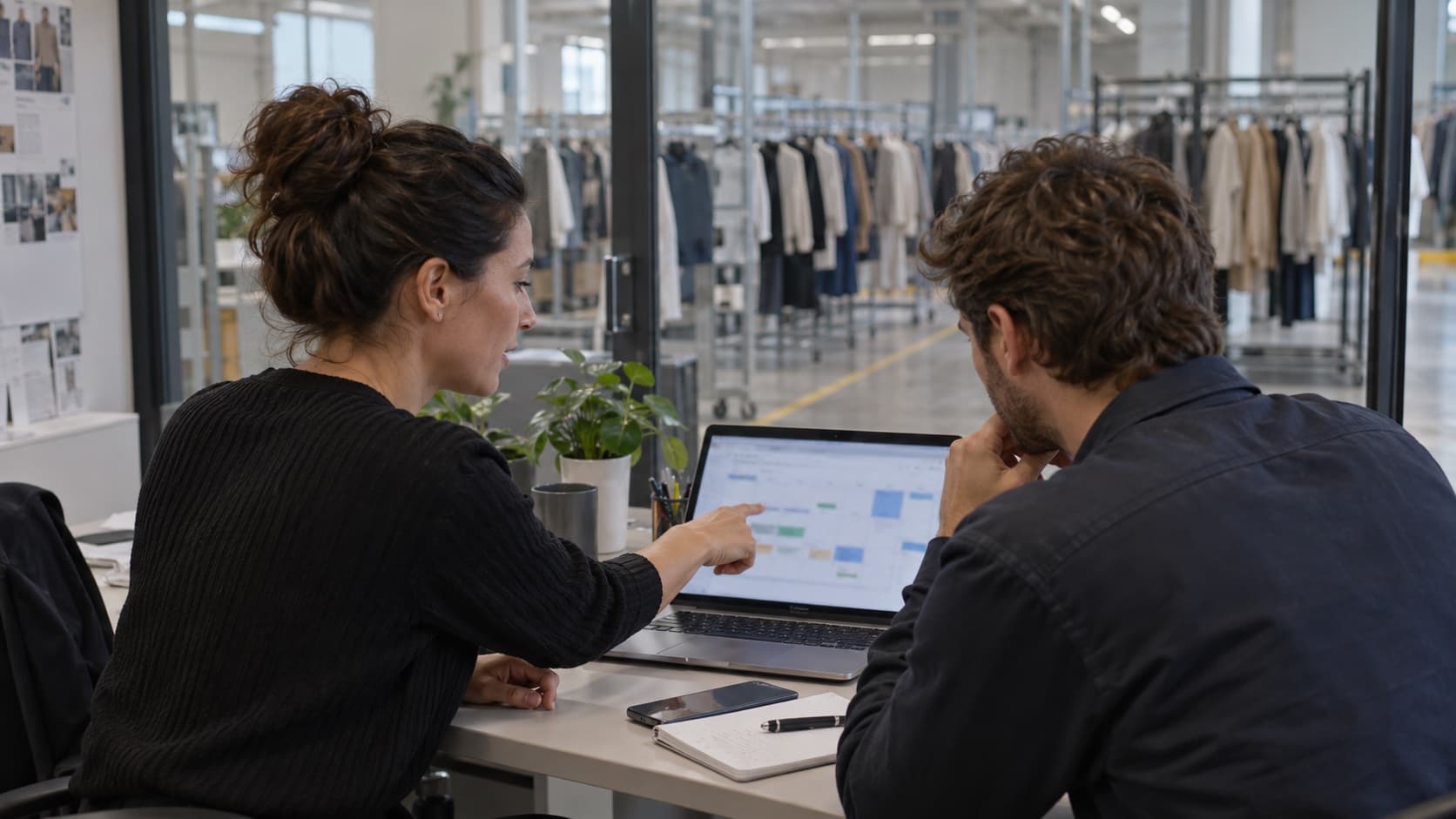Deux professionnels planifient des tournées sur un ordinateur dans un bureau vitré donnant sur un entrepôt textile.